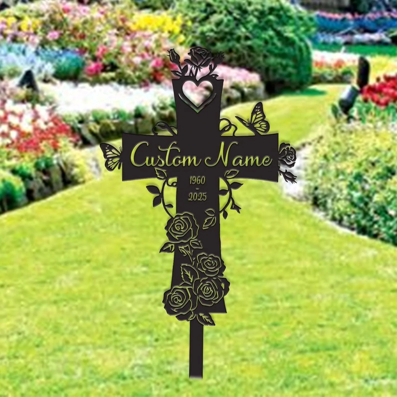 Black metal memorial cross sign with roses, butterflies and placeholder text "Custom Name" and years 1960–2025 staked in a grassy flower garden