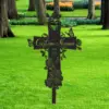 Black metal memorial cross stake with roses, butterflies and placeholder text "Custom Name" and dates, placed in a grassy park with trees