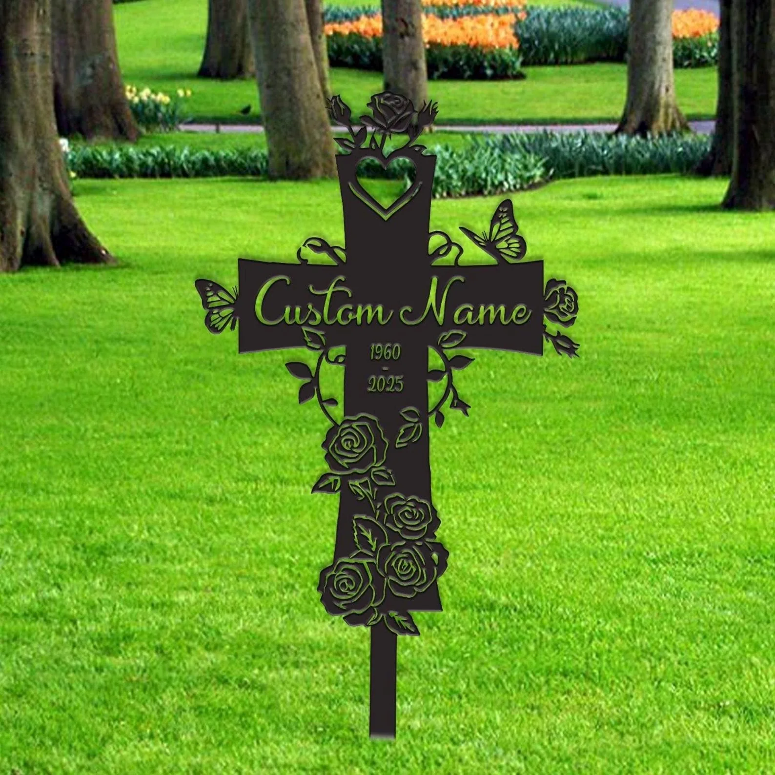 Black metal memorial cross stake with roses, butterflies and placeholder text "Custom Name" and dates, placed in a grassy park with trees