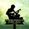 Black silhouette of a seated person playing guitar above a memorial sign reading "In Loving Memory" and space for a custom name and dates against a green grassy background