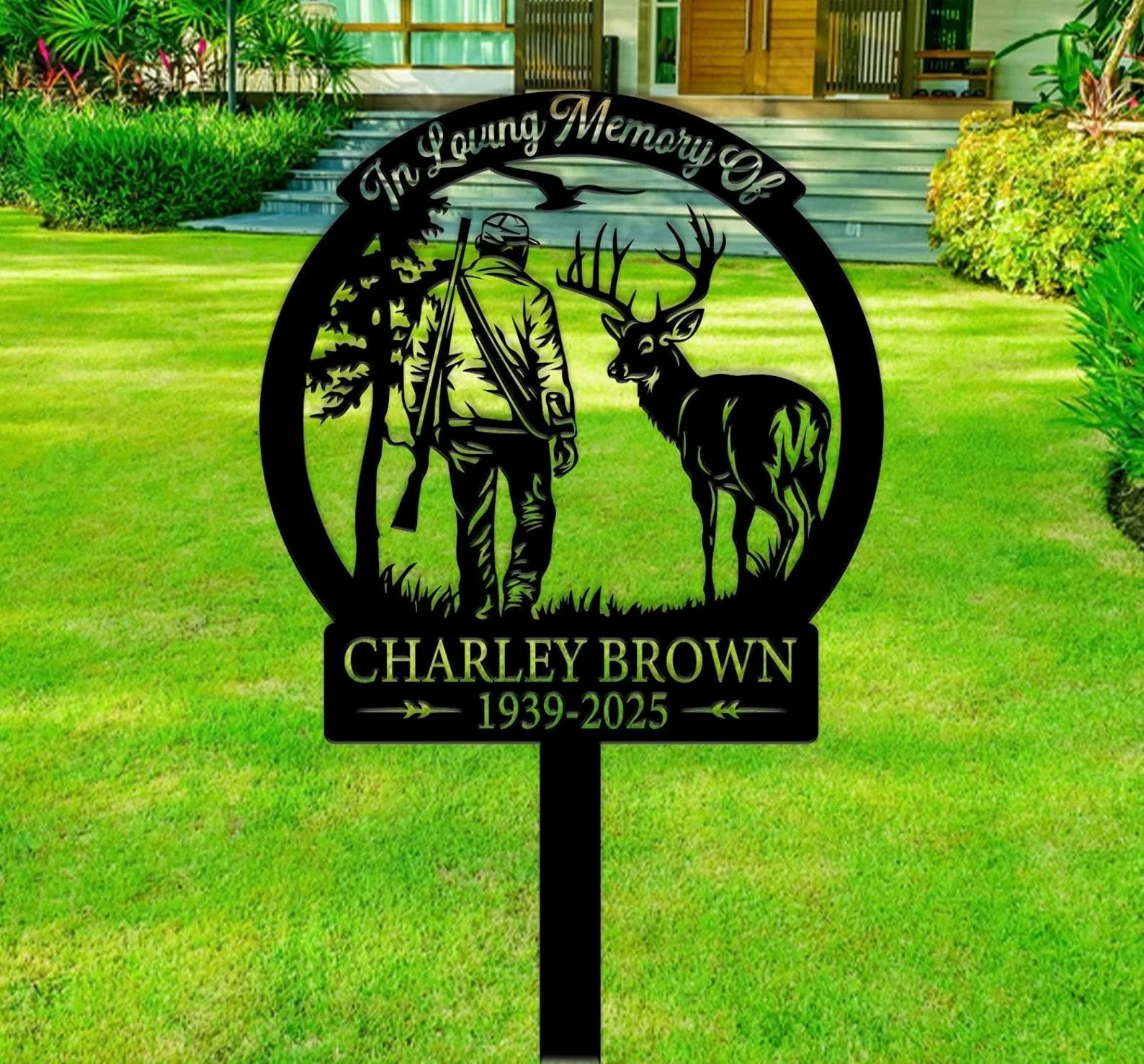 Black metal memorial yard sign in front of a lawn showing a hunter with a rifle and a deer inside a circular frame reading “In Loving Memory Of” and the name “Charley Brown 1939-2025”