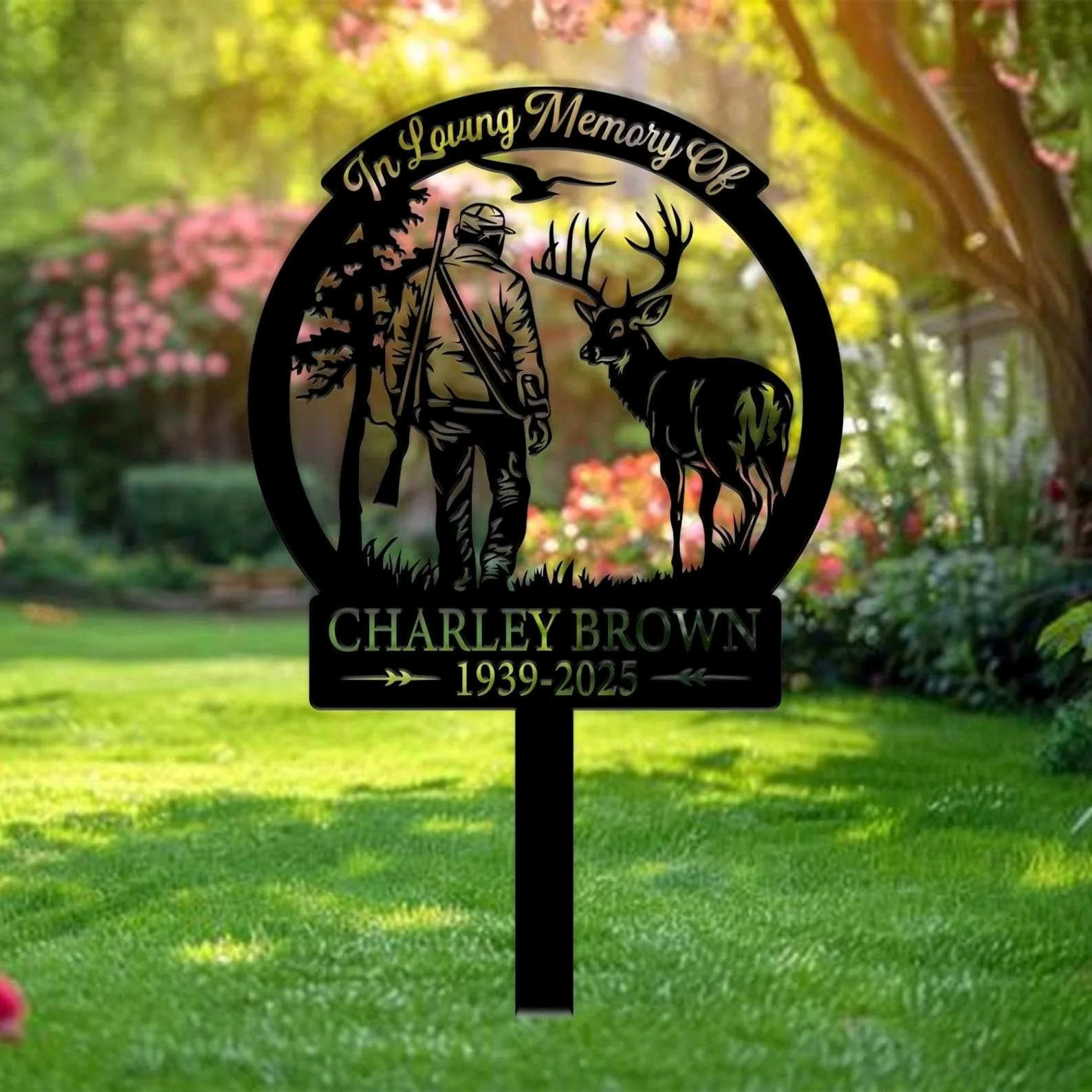 Black metal memorial yard sign reading "In Loving Memory Of" and "Charley Brown 1939-2025" showing a hunter with rifle and a stag silhouette against a garden lawn background
