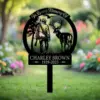 Black metal memorial garden stake reading "In Loving Memory Of" with silhouette of a hunter and deer and name "Charley Brown 1939-2025" against a blurred flower garden background