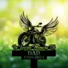 Black silhouette memorial sign of a winged motorcycle reading "In Loving Memory Of" and "DAD 1949-2024" against a sunny grassy background
