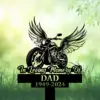 Black memorial sign in grass with winged motorcycle graphic and text reading "In Loving Memory Of DAD 1949-2024" against a soft green bokeh background