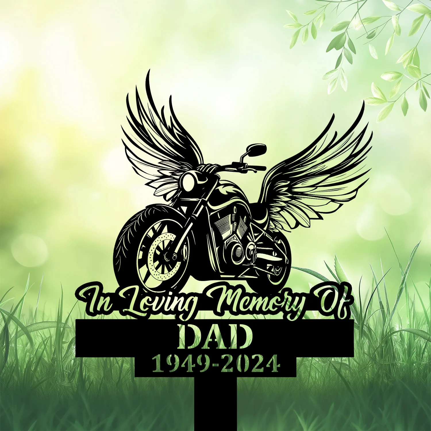 Black memorial sign in grass with winged motorcycle graphic and text reading "In Loving Memory Of DAD 1949-2024" against a soft green bokeh background