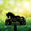 Black silhouette of a horse lying in grass with floral cutouts, name "Stella" and dates "2012-2023" on a garden memorial stake against a sunlit green bokeh background