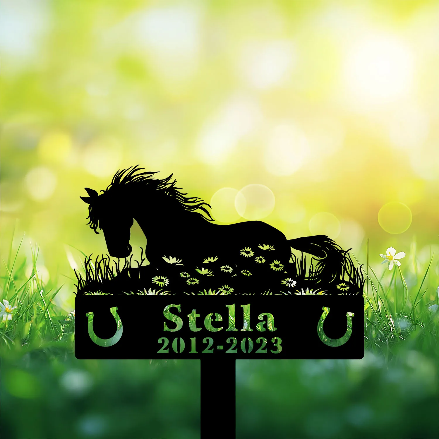 Black silhouette of a horse lying in grass with floral cutouts, name "Stella" and dates "2012-2023" on a garden memorial stake against a sunlit green bokeh background