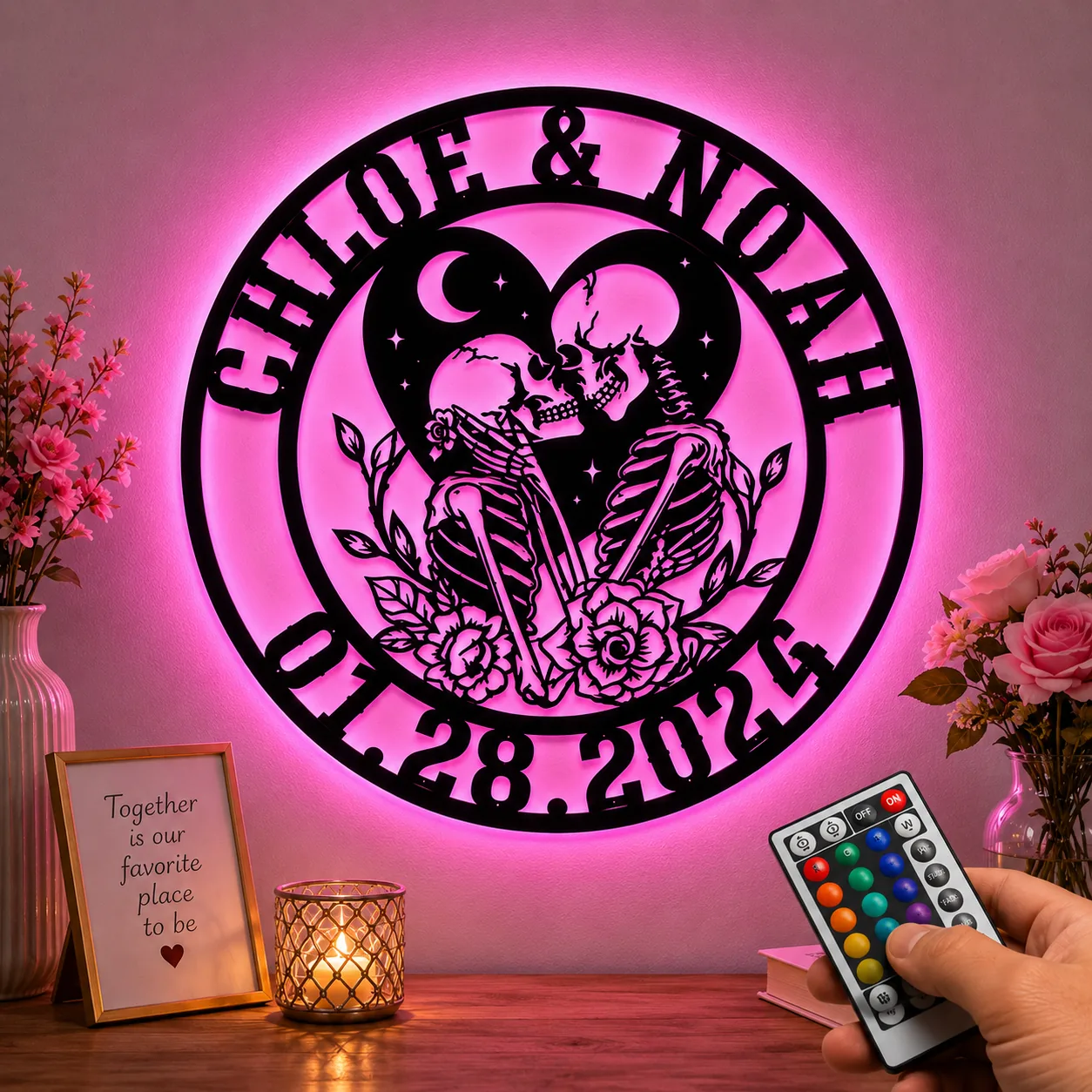 fe5bf4b0-6a48-4bdb-bb9c-c0af97b8679b Round LED wall sign with two kissing skeletons in a heart, names “CHLOE & NOAH” and date “01.28.2024,” glowing pink above a wooden table with flowers, candle, and remote control