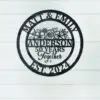 Black round metal sign on a white shiplap wall reading "Matt & Emily" at top, floral design and "ANDERSON 50 YEARS Together" in center, "EST. 2024" at bottom
