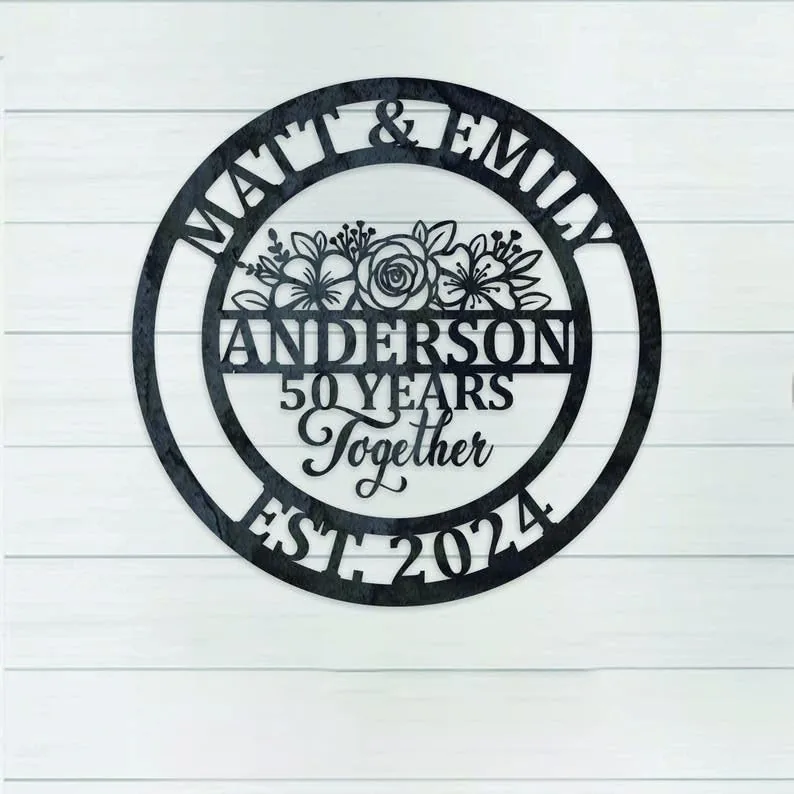 Black round metal sign on a white shiplap wall reading "Matt & Emily" at top, floral design and "ANDERSON 50 YEARS Together" in center, "EST. 2024" at bottom