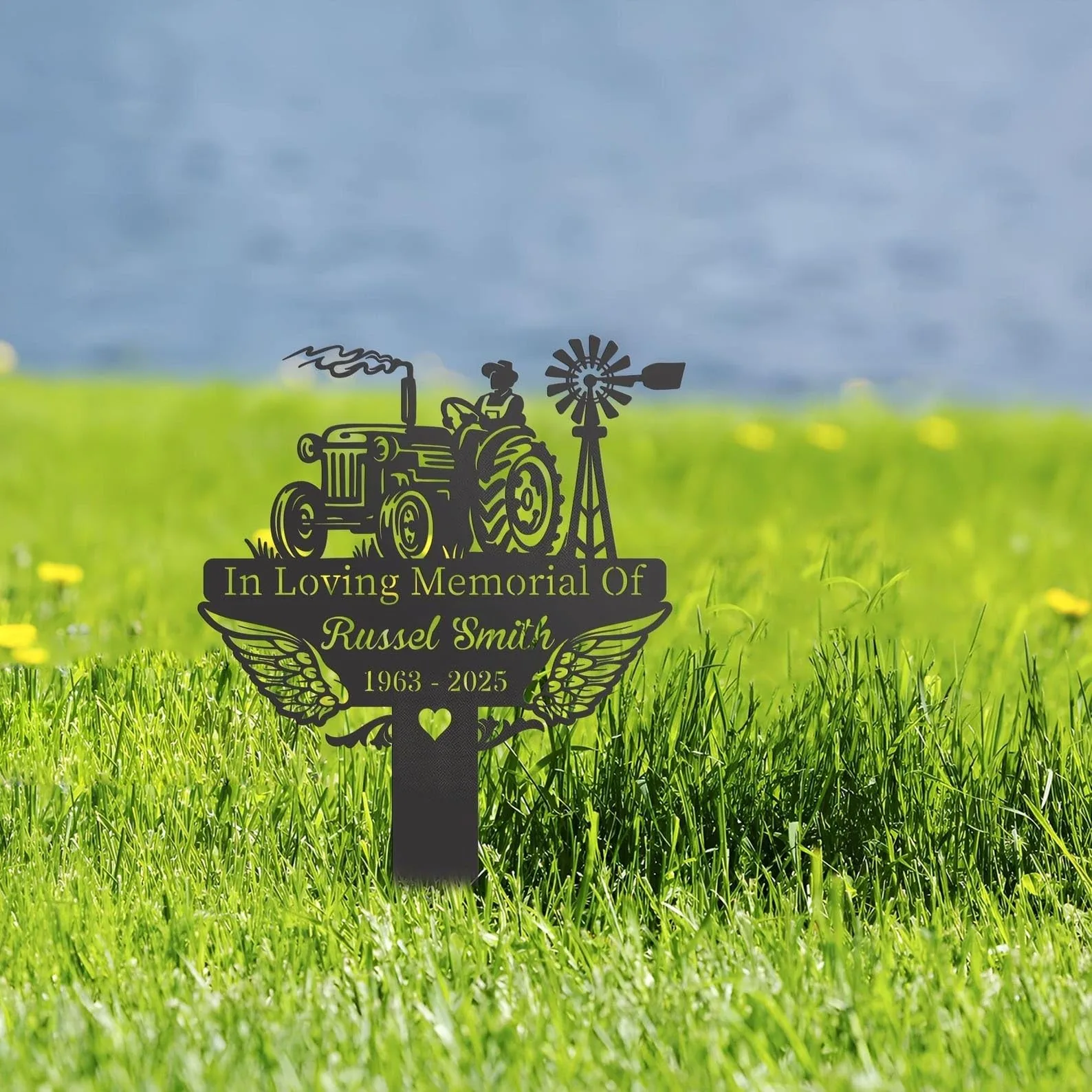 Black metal memorial sign with a tractor and windmill silhouette reading "In Loving Memorial Of Russel Smith 1963 - 2025" staked in a grassy field