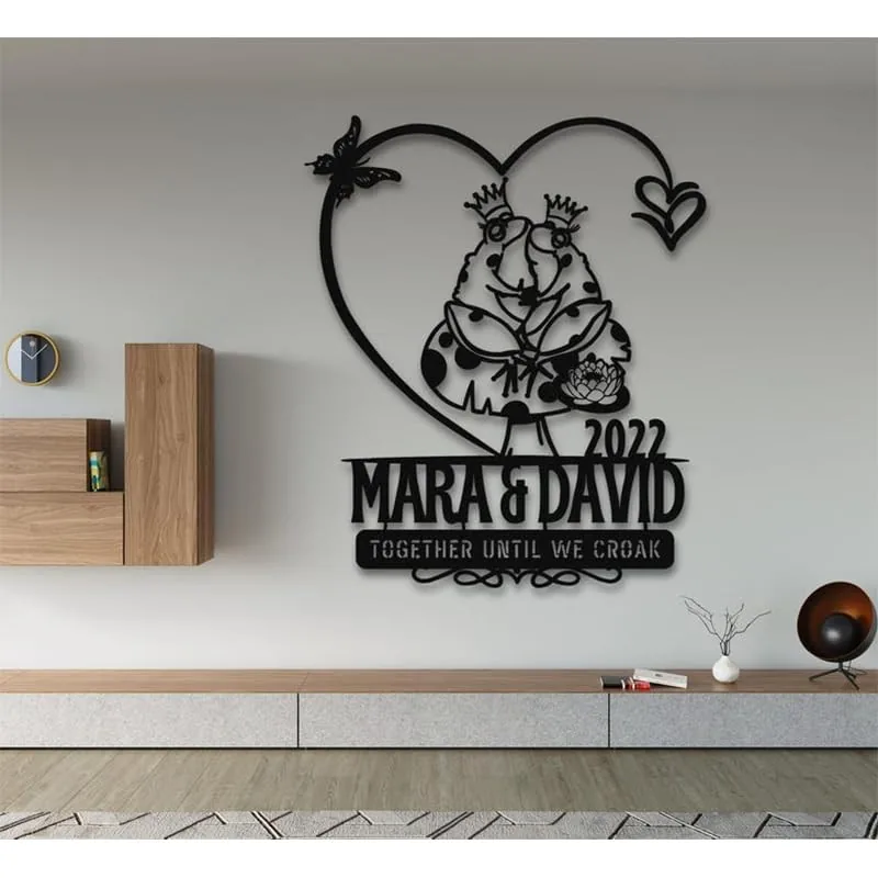 Black metal wall art of two crowned frogs inside a heart above text 'Mara & David 2022' and slogan 'Together Until We Croak' mounted on a modern living room wall
