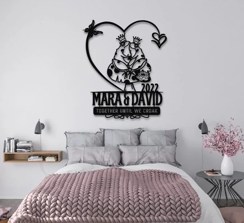 Black metal wall art above bed reading 'MARA & DAVID 2022' with frog couple inside a heart and text 'TOGETHER UNTIL WE CROAK' in modern bedroom