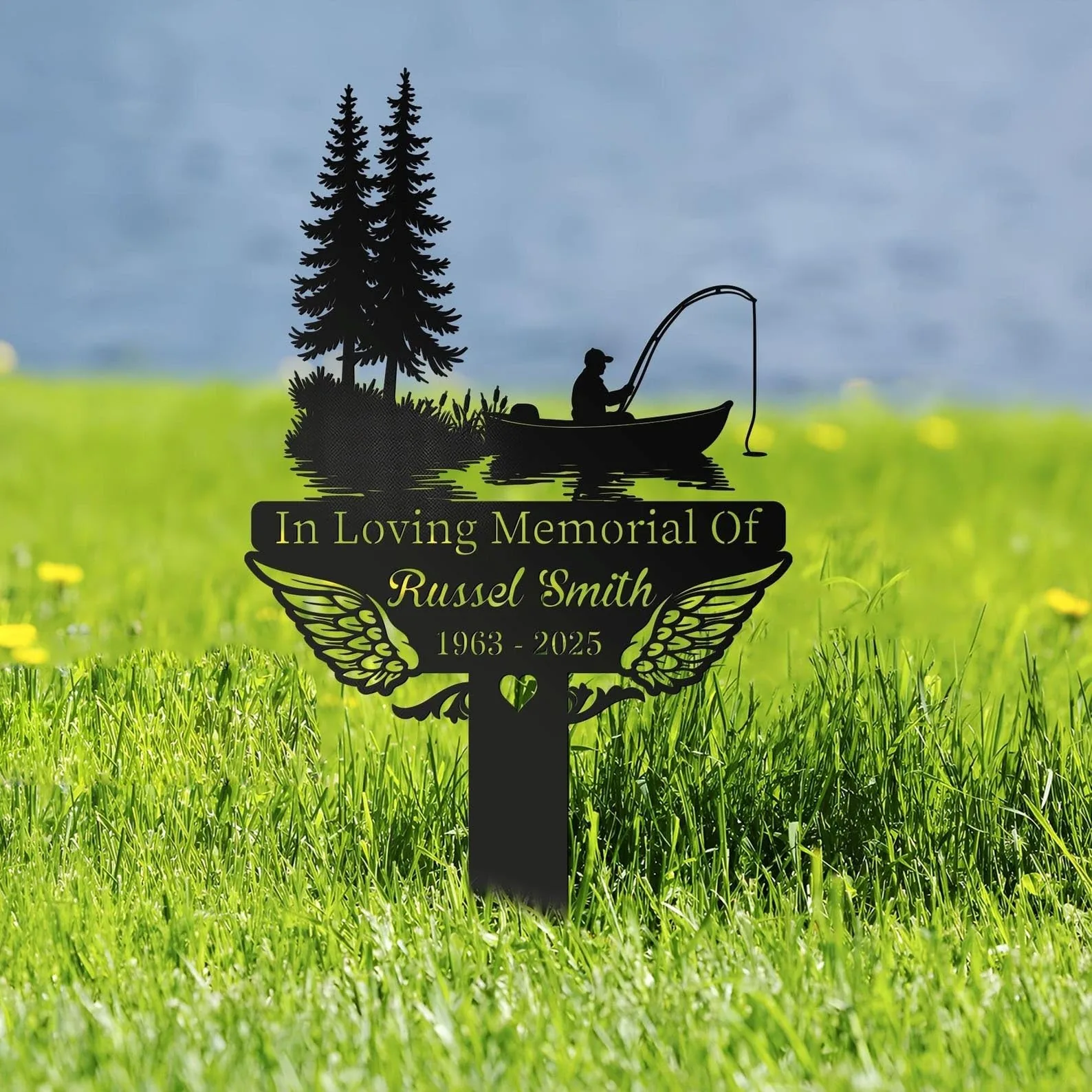 Black metal memorial stake in grass with canoe fishing scene, pine trees, angel wings and the name "Russel Smith" with dates 1963 - 2025