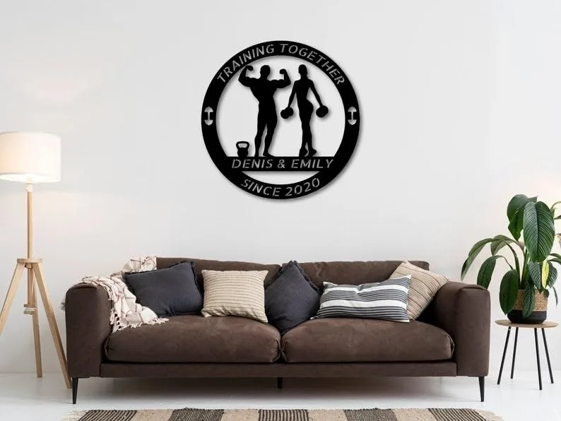 Black circular metal wall sign reading 'Training Together Denis & Emily Since 2020' with silhouettes of a man and woman holding weights, hung above a brown sofa in a modern living room