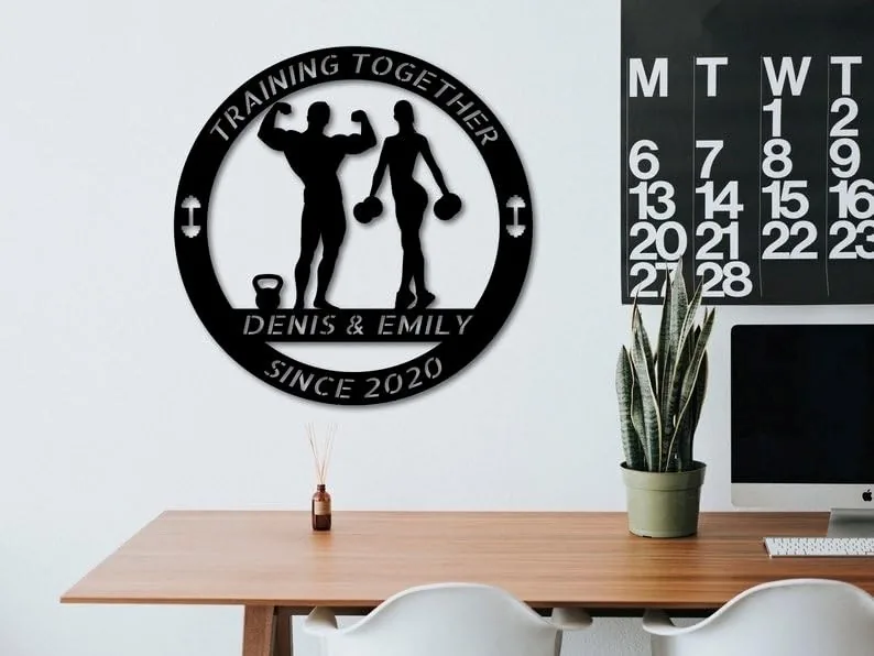 Black round metal wall sign reading 'Training Together' and 'Denis & Emily Since 2020' with silhouettes of a man flexing and a woman holding dumbbells, hung above a desk