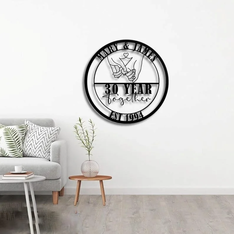 Black round metal wall sign reading “Mary & James” at top, illustration of linked hands, “30 Year Together” and “Est 1994,” hung on a white living room wall above sofa