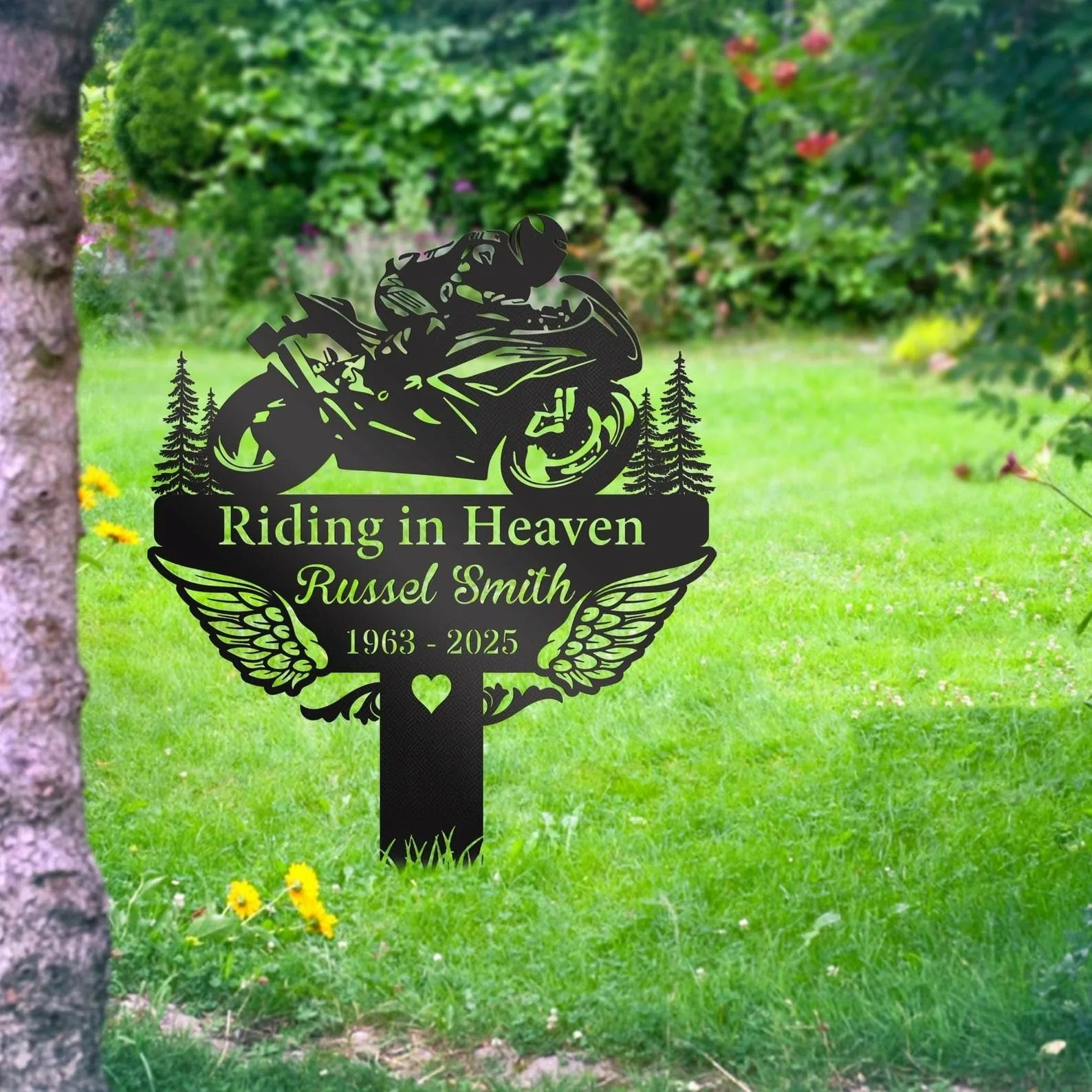 style-17.-Riding-in-Heaven-1 Black metal memorial sign in a grassy garden reading "Riding in Heaven" with name Russel Smith and years 1963–2025, motorcycle silhouette and wings