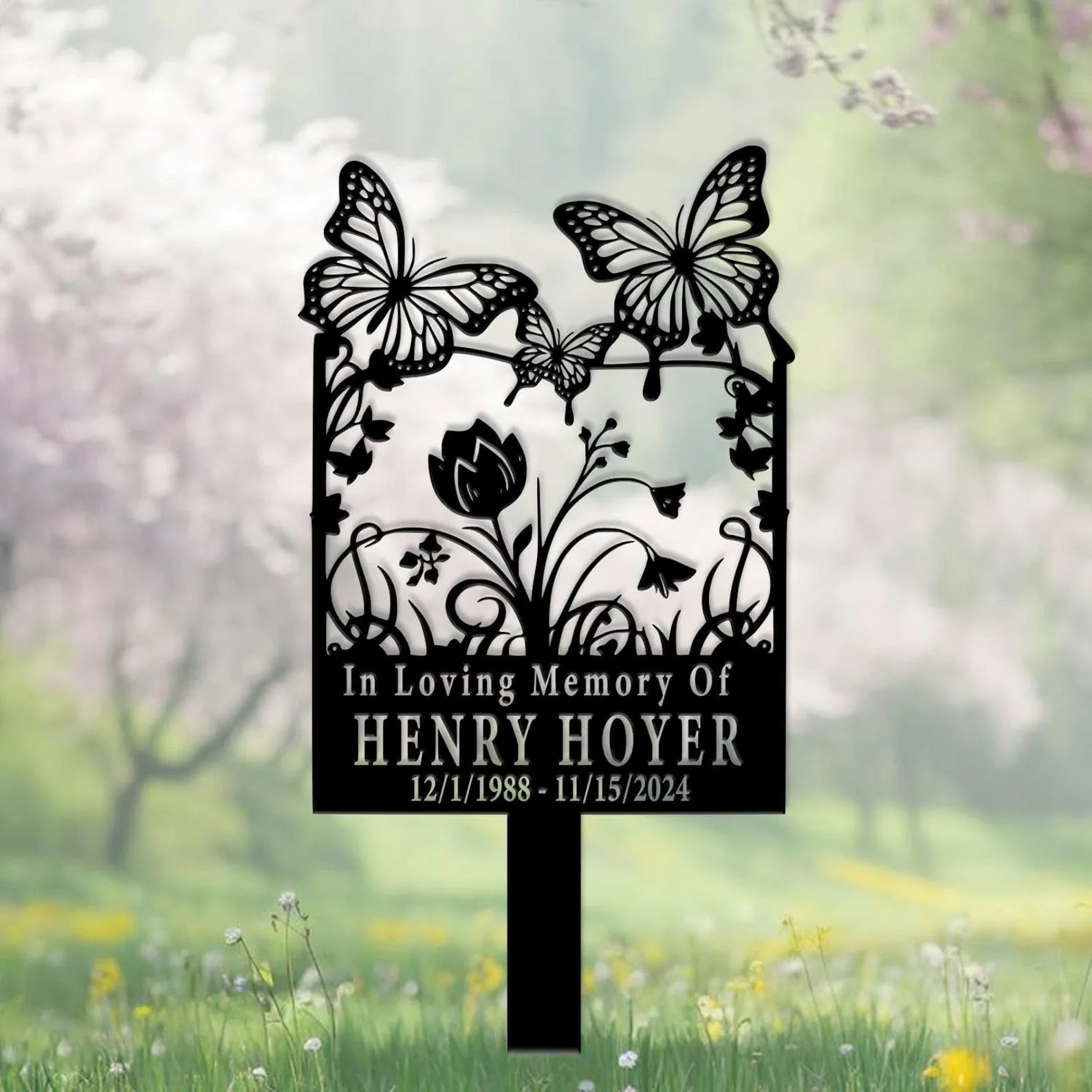 Black metal memorial garden marker with floral and butterfly cutouts reading "In Loving Memory Of HENRY HOYER 12/1/1988 - 11/15/2024" placed in a grassy meadow