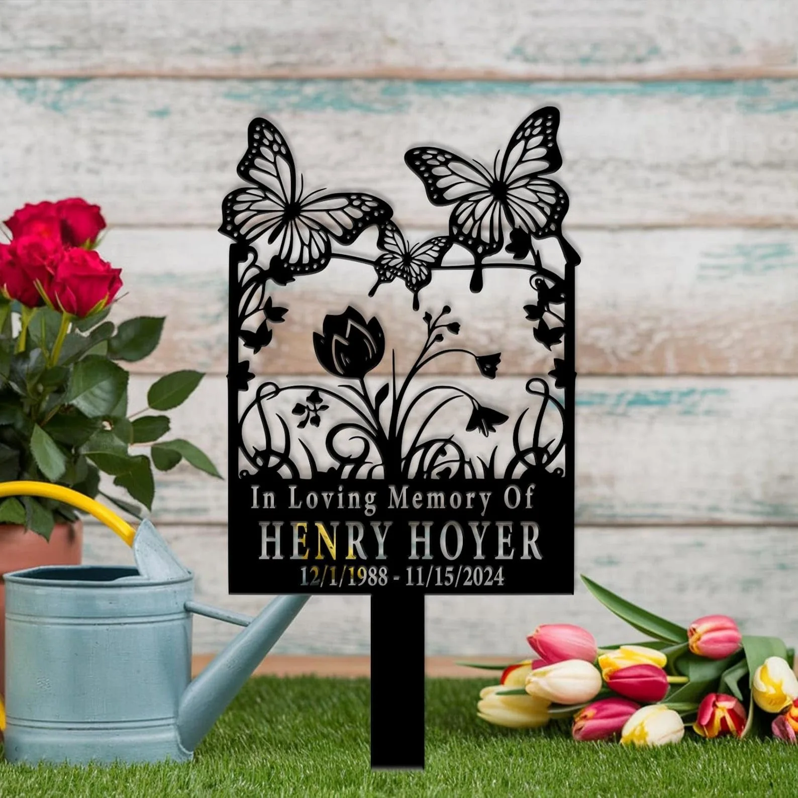 Black metal memorial garden stake with butterflies and flowers reading "In Loving Memory Of HENRY HOYER 12/1/1988 - 11/15/2024" placed on grass with watering can, roses and tulips nearby
