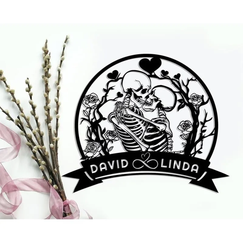 Black cutout wall art of two embracing skeletons surrounded by roses and vines, circular frame with heart motifs and banner reading "David ♥ Linda", displayed on a white background next to pussy willow branches and pink ribbon.