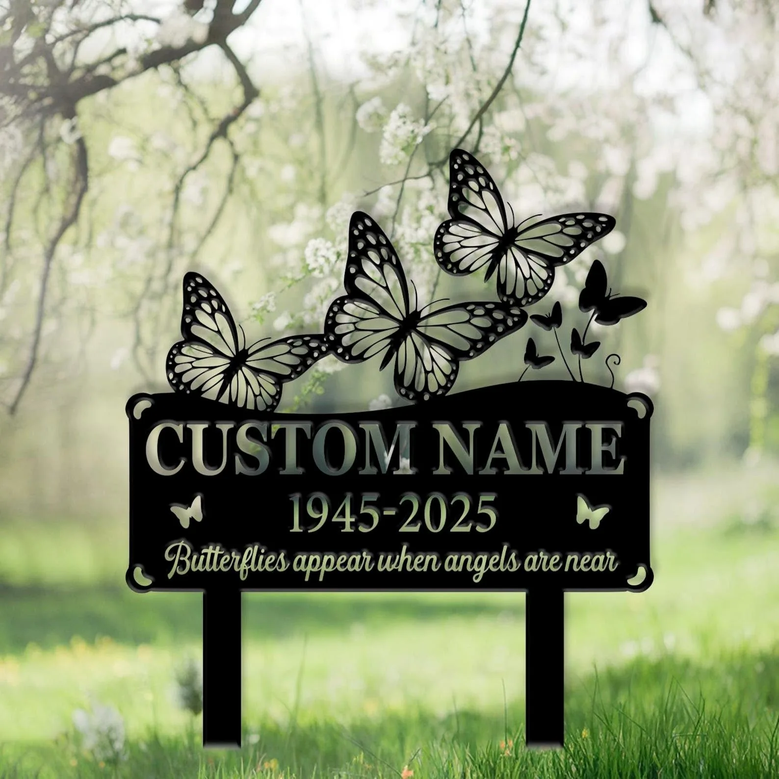 Black metal memorial yard sign with decorative butterflies, placeholders reading "CUSTOM NAME" and dates "1945-2025" against a soft-focused spring garden background
