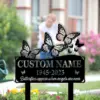 Black memorial garden sign with decorative butterflies and text placeholder "CUSTOM NAME 1945-2025" placed among flowering plants in a front yard