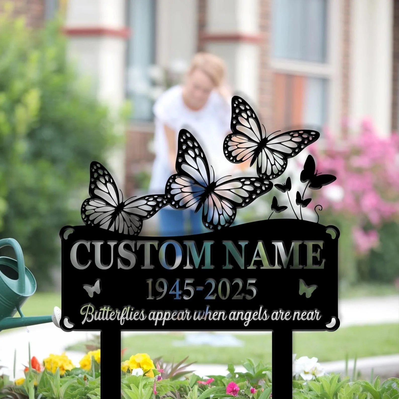 Black memorial garden sign with decorative butterflies and text placeholder "CUSTOM NAME 1945-2025" placed among flowering plants in a front yard