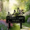 Black metal memorial yard sign with butterfly cutouts and placeholder text showing years 1945-2025, placed in a sunlit garden path among lavender and green foliage