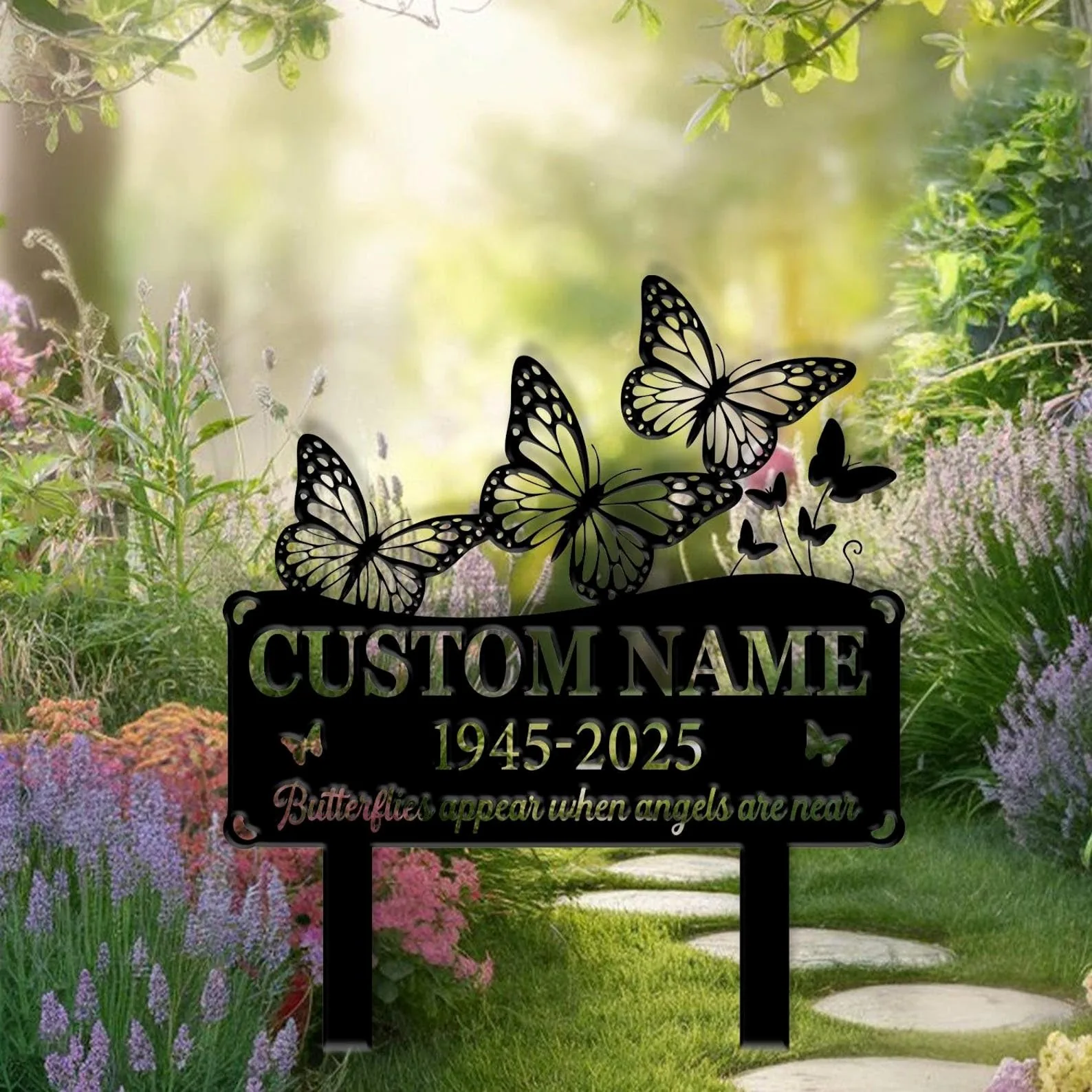 Black metal memorial yard sign with butterfly cutouts and placeholder text showing years 1945-2025, placed in a sunlit garden path among lavender and green foliage