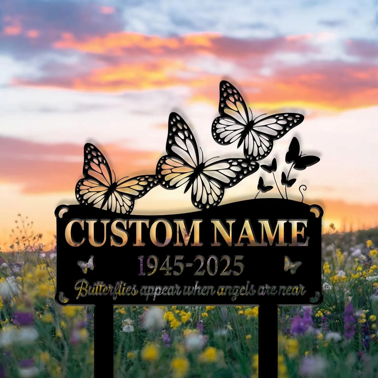 Black memorial sign with cutout butterflies and text placeholder "CUSTOM NAME" and dates "1945-2025" set against a sunset sky over a wildflower field