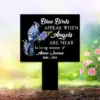 Black memorial garden marker with a blue bird and purple flowers reading "Blue Birds appear when Angels are near. In loving memory of Anna James 1956-2022," placed in a sunny garden near pink daisies.