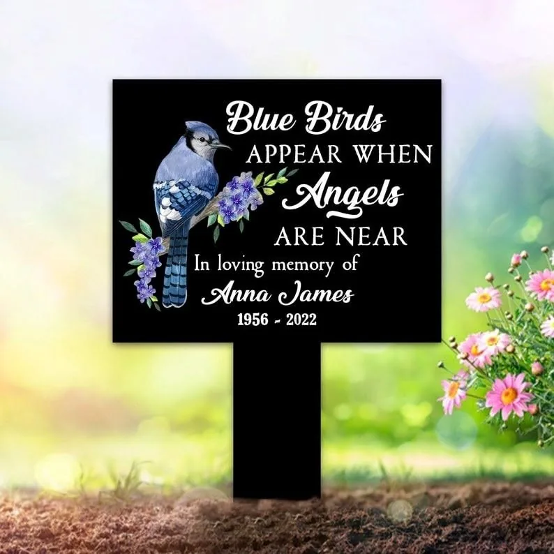 Black memorial garden marker with a blue bird and purple flowers reading "Blue Birds appear when Angels are near. In loving memory of Anna James 1956-2022," placed in a sunny garden near pink daisies.