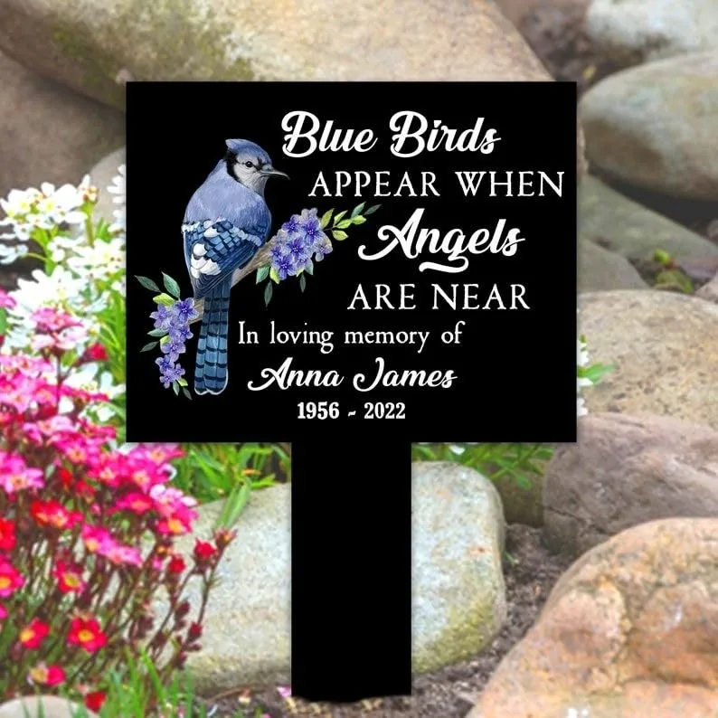 Black memorial garden stake with a blue bird and purple flowers reading “Blue Birds appear when Angels are near In loving memory of Anna James 1956 - 2022,” placed among rocks and pink garden flowers