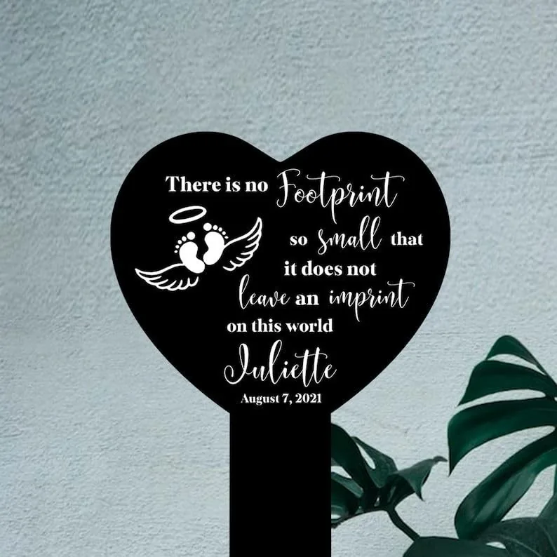 Black heart-shaped memorial sign with white text reading a remembrance quote, baby footprints with wings and halo, name "Juliette" and date August 7, 2021, displayed against a textured light wall near a plant.