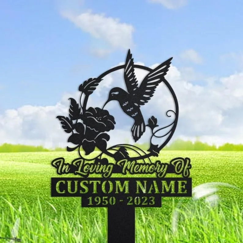 Black metal memorial yard sign with a hummingbird and flower design reading "In Loving Memory Of" and placeholder "CUSTOM NAME 1950 - 2023" against a grassy field