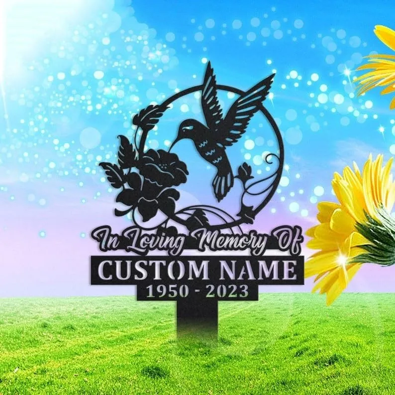 Black metal memorial stake sign reading "In Loving Memory Of" with placeholder "CUSTOM NAME" and dates 1950-2023, hummingbird and flower design on a grassy background