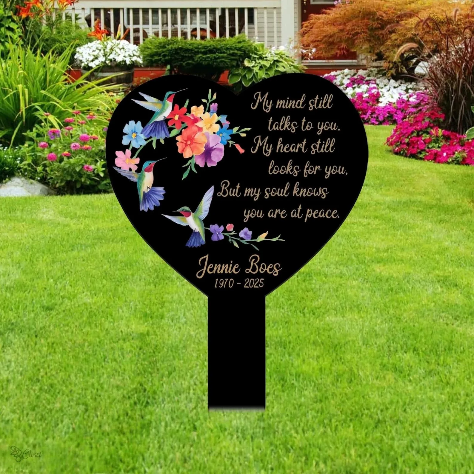 style-25.-Heart-Hummingbirds-1 Black heart memorial yard sign with colorful hummingbirds and flowers, text reading “My mind still talks to you, My heart still looks for you, But my soul knows you are at peace.” and name Jennie Boes 1970-2025, placed in a green front lawn with flower beds