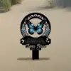 Black memorial garden stake in sand featuring a blue butterfly, floral wreath, poem text and name “Elena Rose 2022 - 2024”