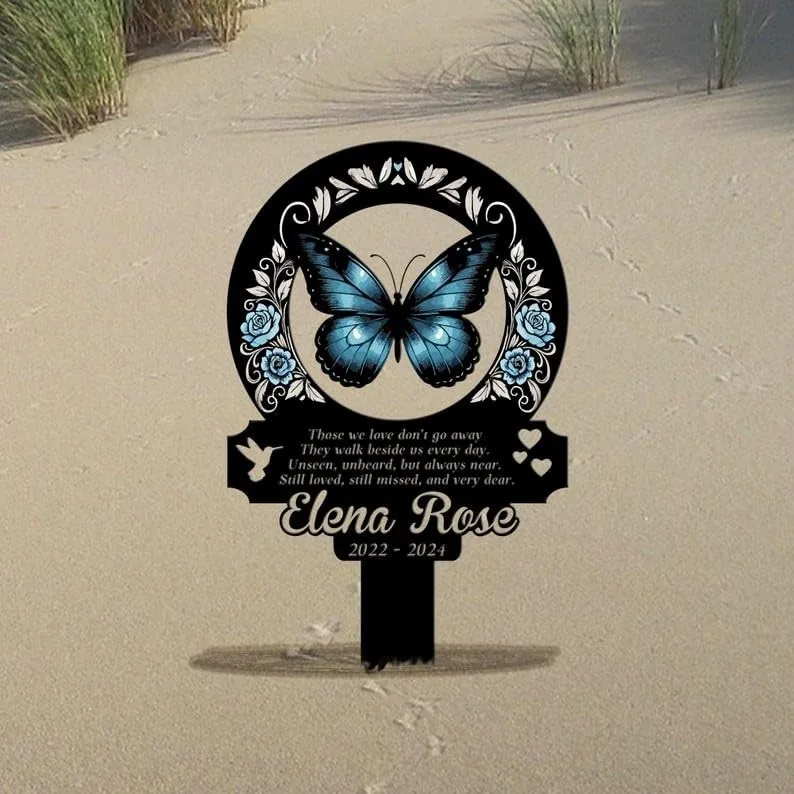 Black memorial garden stake in sand featuring a blue butterfly, floral wreath, poem text and name “Elena Rose 2022 - 2024”