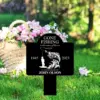 Black memorial garden sign reading “GONE FISHING in the waters of heaven,” years 1945–2023, fishing graphic and “In loving memory of John Olson,” staked in green lawn near a basket of pink flowers