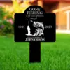 Black memorial yard sign with white fishing design reading 'Gone Fishing in the waters of heaven', years 1945–2023, and 'In loving memory of John Olson' placed in green lawn