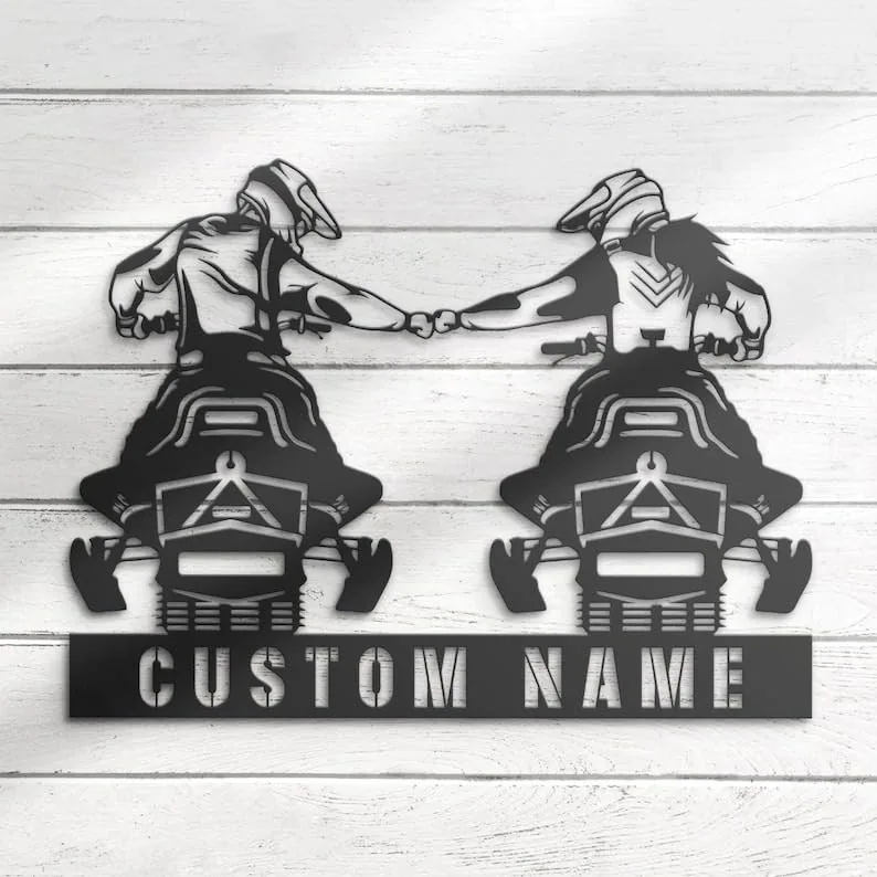Black metal wall art of two snowmobile riders fist-bumping above a rectangular plate reading 'CUSTOM NAME', mounted on white wooden planks