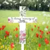 White memorial cross with floral accents reading "In loving memory of Emily 1963-2024" placed in a wildflower meadow with red poppies and green trees in background