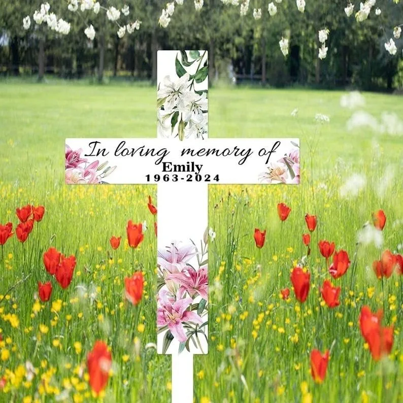 White memorial cross with floral accents reading "In loving memory of Emily 1963-2024" placed in a wildflower meadow with red poppies and green trees in background
