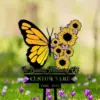 Yellow and black butterfly shape made of sunflowers and small pink butterflies above text reading "In Loving Memory Of" and placeholder "CUSTOM NAME 1988 - 2024" on a stake in a grassy field with purple flowers