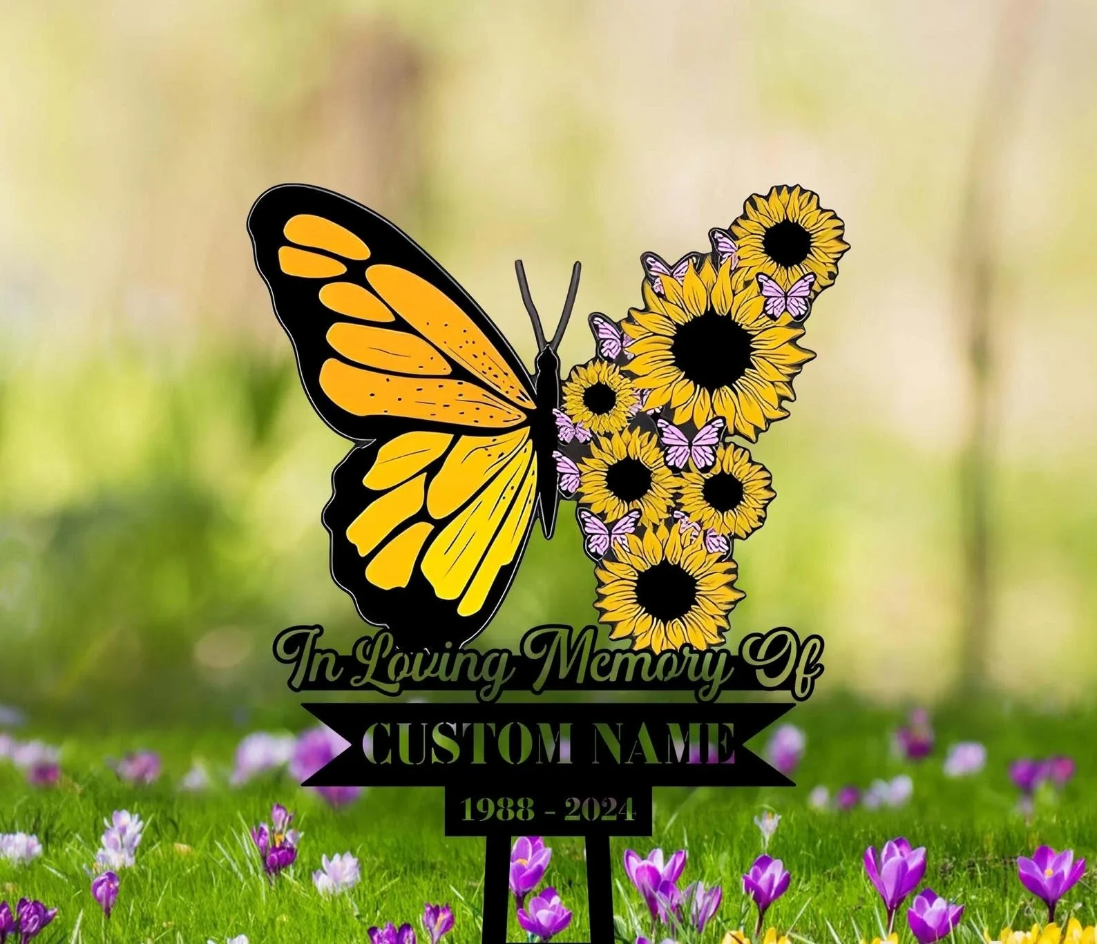 Yellow and black butterfly shape made of sunflowers and small pink butterflies above text reading "In Loving Memory Of" and placeholder "CUSTOM NAME 1988 - 2024" on a stake in a grassy field with purple flowers