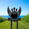 Black metal yard sign of a winged bowling ball and pins with text 'CUSTOM NAME' and dates '1960 - 2022' placed on grassy coastal lawn by the ocean
