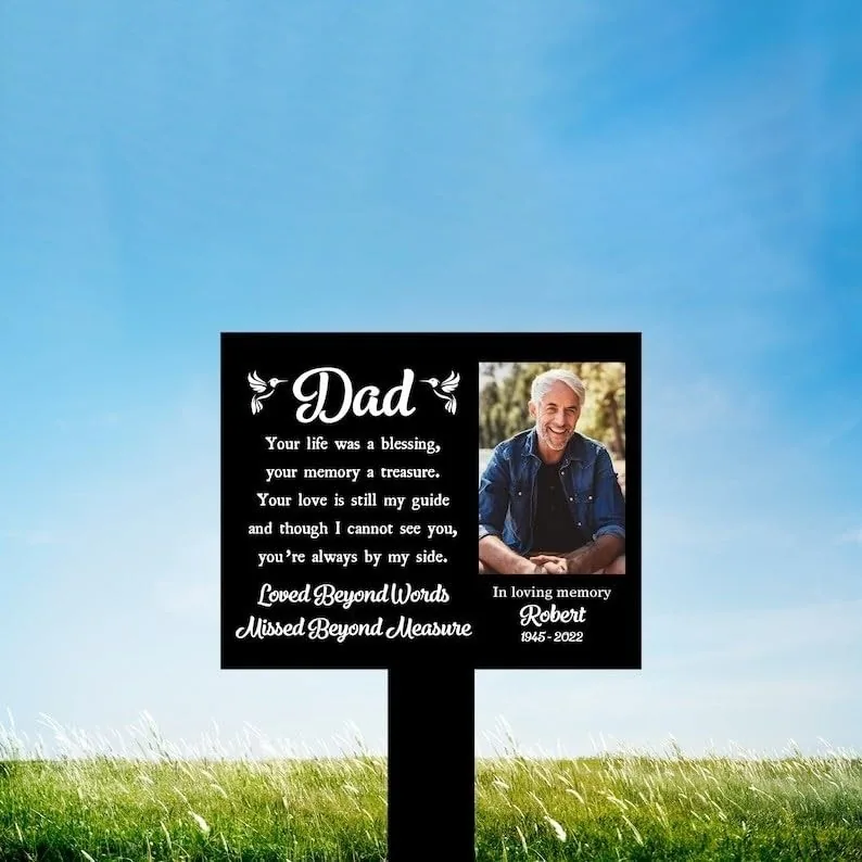 Black memorial lawn sign reading “Dad” with poem and “Loved Beyond Words Missed Beyond Measure,” photo of smiling man and text “In loving memory Robert 1945–2022,” set in grassy field